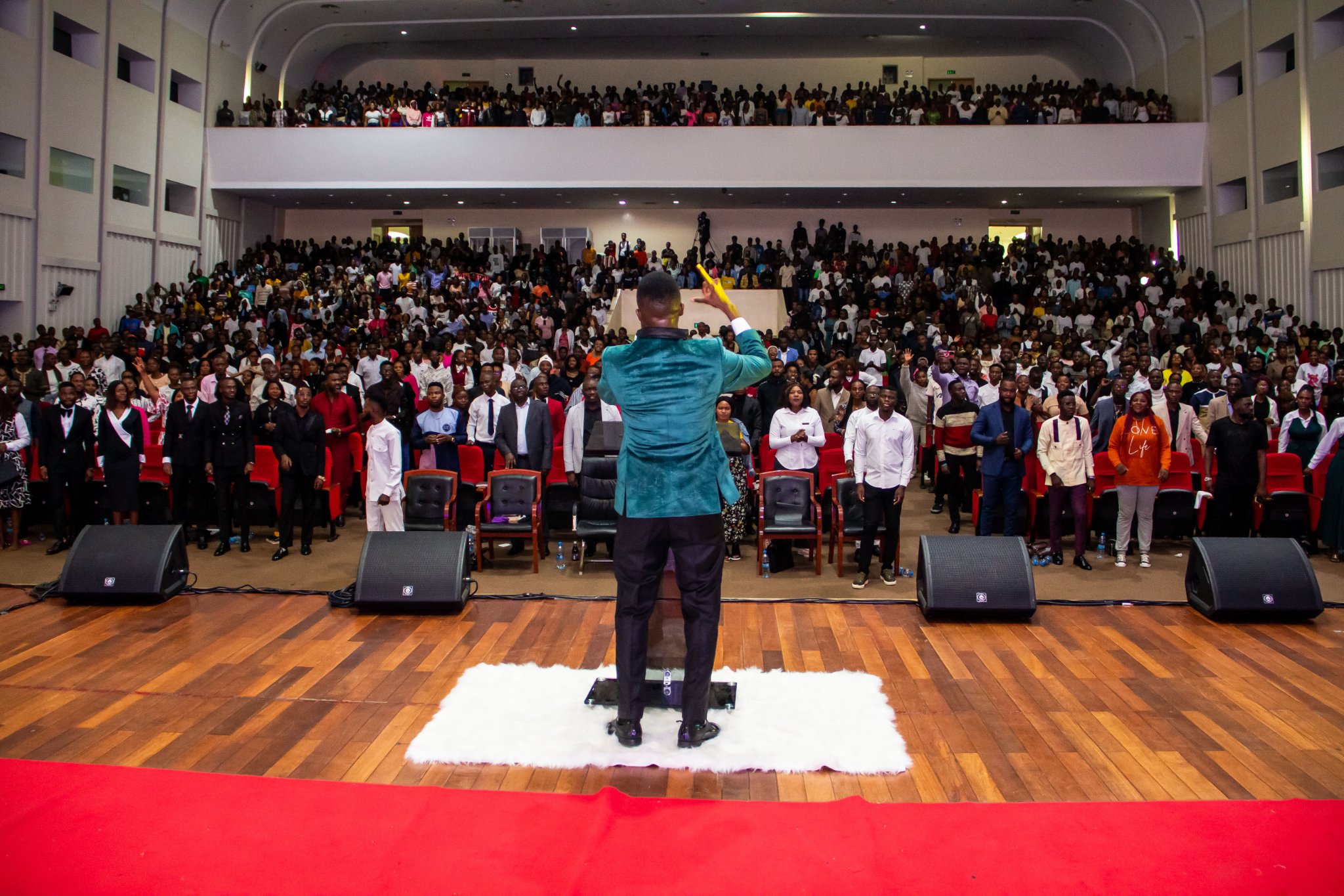 A preacher in a teal jacket seen from behind on stage before a packed auditorium